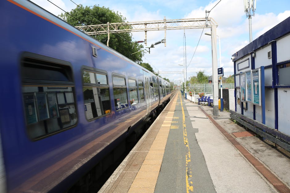 A blue train is stopped at an outdoor station platform, with the side windows reflecting the surrounding environment. The platform features a yellow safety line along the edge, next to a narrow sectio