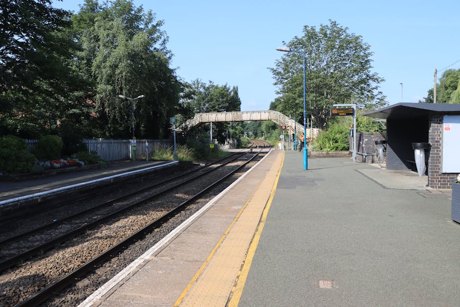 A blue train is stopped at an outdoor station platform, with the side windows reflecting the surrounding environment. The platform features a yellow safety line along the edge, next to a narrow sectio
