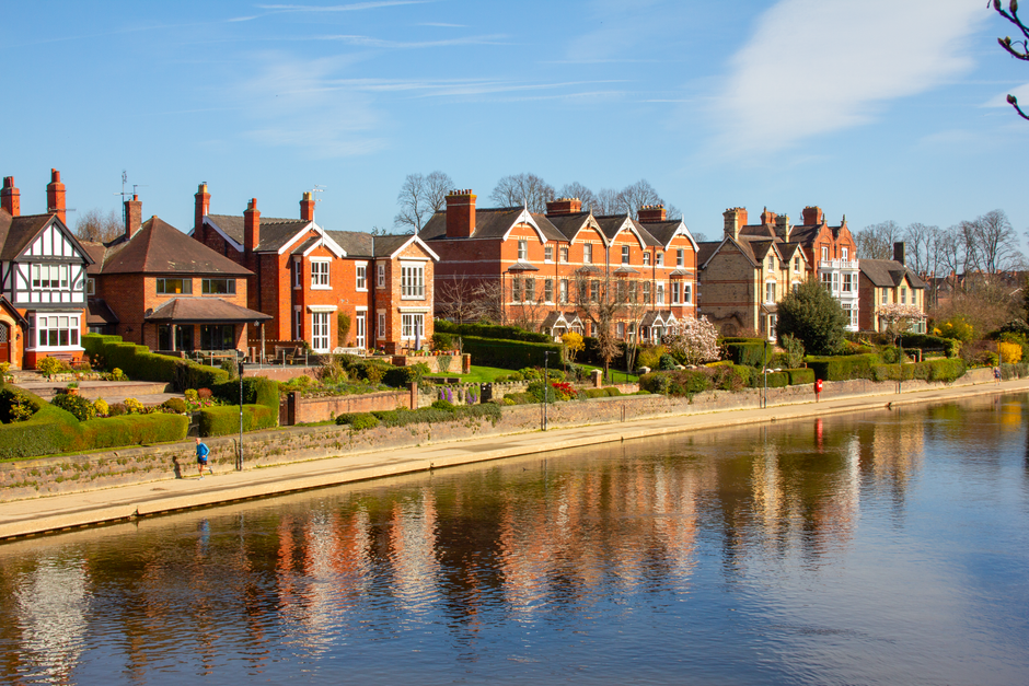 A row of three red-brick suburban houses along a narrow canal with clear, gently flowing water. The houses feature pitched roofs with dark and grey tiles, white-framed windows, and small front gardens